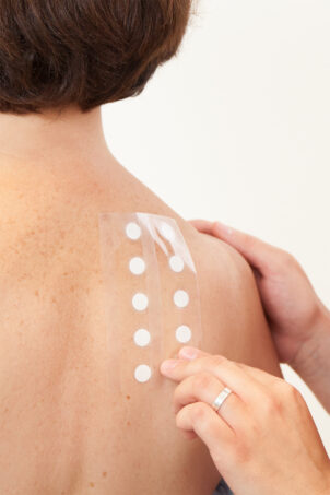 Image of a woman from behind with path tests applied by an external hand, symbol of the blog article dedicated to clinical tests in makeup: a competitive advantage because they are fundamental for beauty brands, of the cosmetic & medical device business unit of Abich_Lifeanalytics